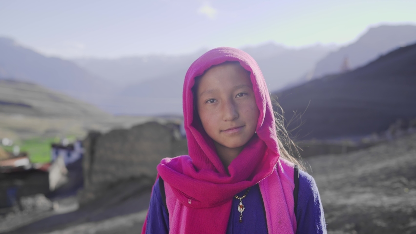 A happy young rural Asian Indian girl with a colorful scarf around her head and neck standing against morning sunlight with the mountain rural village in the back looking at the camera and smiling Royalty-Free Stock Footage #1081445774