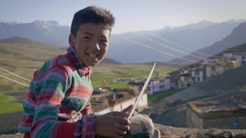 A cheerful young Asian rural boy sitting outdoors in the morning sunlight with a laptop smiling looking at a camera with a landscape view of a mountainous region with a tiny village on its foothills. Royalty-Free Stock Footage #1081445777