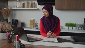 Friendly lovely muslim freelance female in hijab wearing hands free headset, communicating and providing customer online support, checking available merchandise while working at home.