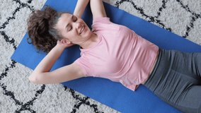 Home training and active lifestyle. Pretty woman with curly hair in pink t-shirt does abdominal crunches and rests on floor with carpet in light room upper view 4k video