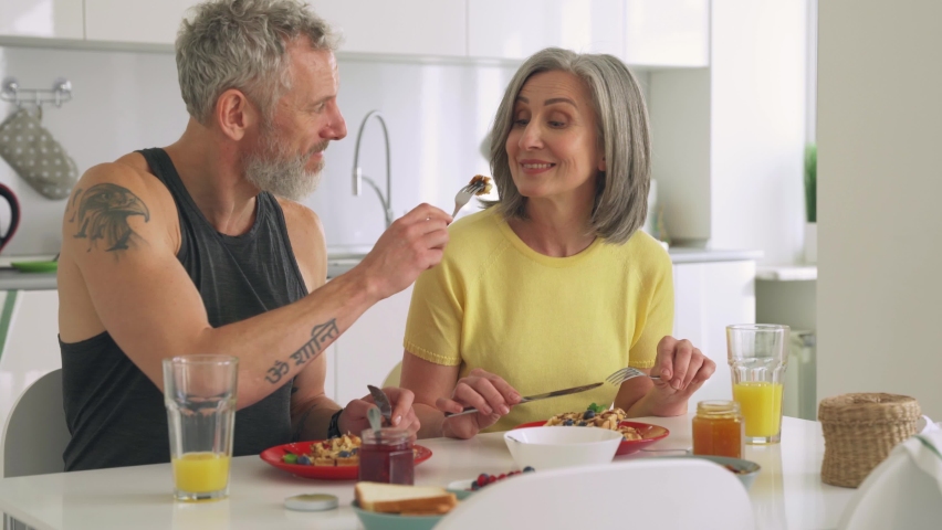 Happy healthy mature older family having breakfast at home. Husband feeding wife tasting waffles. Affectionate senior couple eating dessert sitting at kitchen embracing enjoying morning meal together. Royalty-Free Stock Footage #1081453505