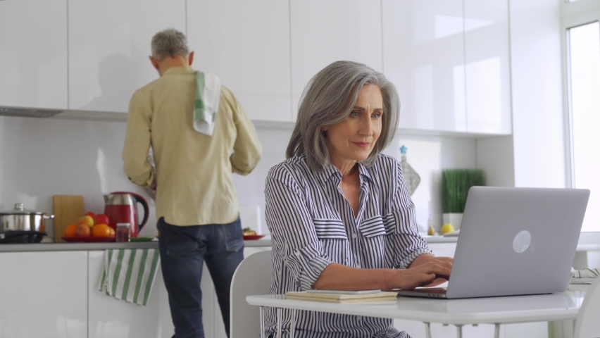 Mature mid aged 50s woman wife remote working or distance learning online from home office using laptop cellphone while her senior husband cooking breakfast in kitchen helping with household work. | Shutterstock HD Video #1081453511