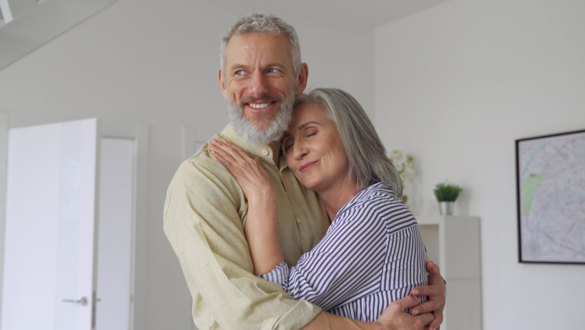 Dreamy senior adult mature couple standing hugging, bonding, thinking of good future. Carefree cheerful mid age old husband hugging wife looking away dreaming, enjoying wellbeing love in new house. | Shutterstock HD Video #1081453526