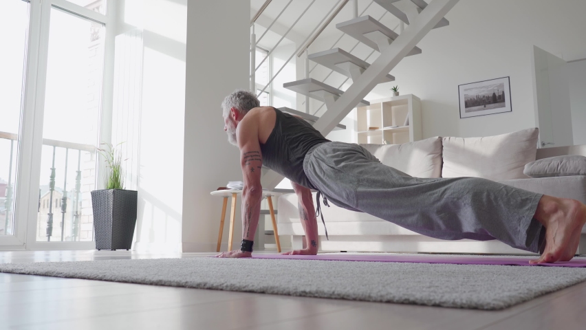 Fit sporty active tattooed senior mature mid age hipster man doing morning workout exercise at home, enjoying sport pilates yoga fitness training on mat in modern living room.