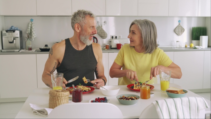 Healthy happy fit mature older family couple having breakfast sitting at kitchen table. Smiling mid age 50s husband and wife talking while eating waffles, having fun enjoying morning meal together. | Shutterstock HD Video #1081453559