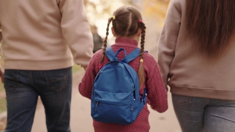 mother and father hold a little schoolgirl with a backpack by the hand, happy family, walk through schoolyard in fall, mom, dad and kid with bag over their shoulders, accompany child to first grade