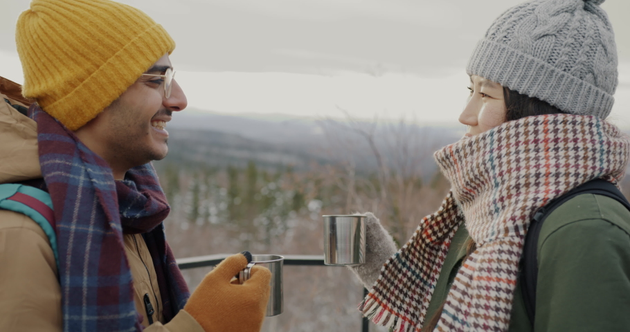 Happy couple tourists clinking metal cups drinking laughing and watching amazing winter landscape mountains and forest. People and tourism concept.