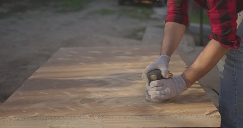A man in a respirator, goggles, polishes a parquet board with an orbital sander.