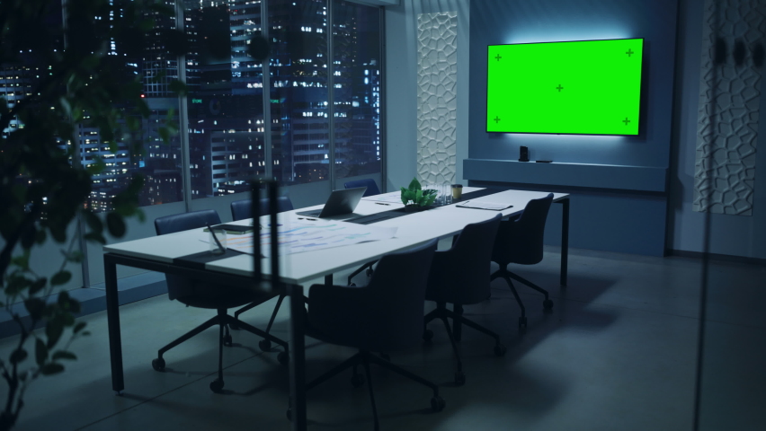 Modern Empty Meeting Room with Big Conference Table with Various Documents and Laptop on it. Wall TV Showing Green Chroma Key Screen Mock Up Display Template. Late Evening or Night City Outside. Royalty-Free Stock Footage #1081459277
