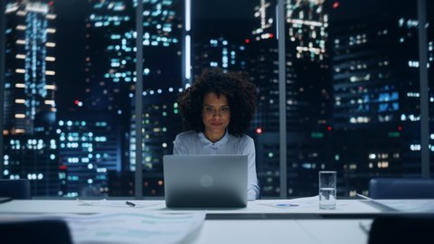 Portrait of a Successful African American Businesswoman Working on Laptop Computer in Big City Office Late in the Evening. Happy Female with Afro Hair in Stylish Blue Blouse Smiles for Camera.