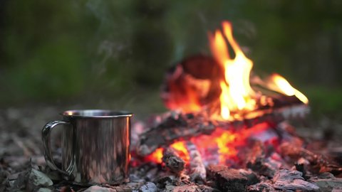 A metal mug with a drink stands next to the fire. Picnic in a camp in the woods. Fireplace in the camp, twilight