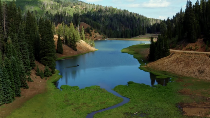 Beautiful ascending aerial drone shot of a stunning nature landscape of the Anderson Meadow Reservoir lake up Beaver Canyon in Utah with large pine tree forest, a small stream, and a grass field.