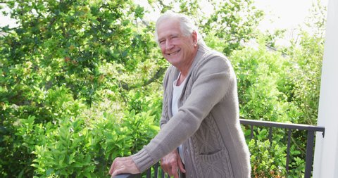 Portrait of smiling retired senior man wearing cardigan sweater leaning on railing in balcony. senior domestic lifestyle.
