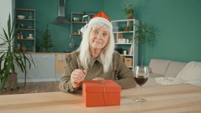A Happy Elderly Woman Wearing Santa Claus Hat Celebrates Christmas Online Enjoy Conversation Making Video Call. A Smiling Woman Enthusiastically Opens Gift Box While Chatting. Celebration Online.