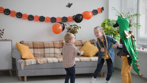 cheerful active children are celebrating halloween party, cheerful boy in dragon costume is playing with sister and brother in game catch me in room decorated with balls and garlands with pumpkin