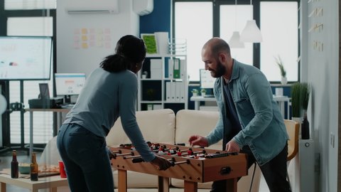 Diverse group of workmates enjoying game with foosball table to do fun activity after work. Coworkers playing football and drinking beer from bottles, celebrating party with entertainment