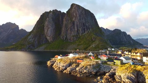 Drone flying over small fishing village Hamnøy in Norway towards iconic mountains above the sea with wooden drying racks for torrfisk