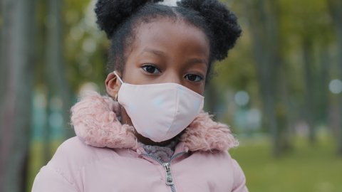 Close up kid face little girl in medical mask looks away turning head looking at camera in city street. Portrait of masked child baby pupil schoolgirl posing in park pandemic coronavirus quarantine