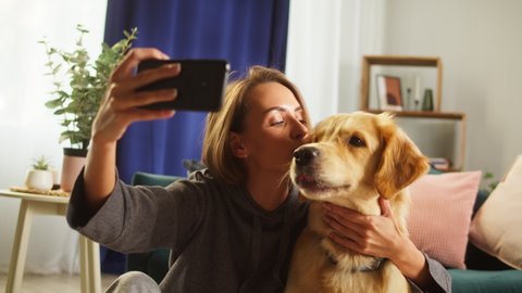 Young woman making selfie with dog in living-room, animal trainer taking photo with golden retriever, using smartphone. Having fun together with lovely pet. Happy puppy labrador posing. 