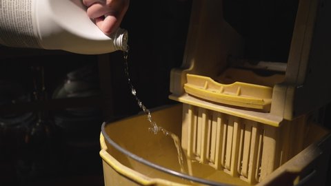 This video shows a hand pouring bleach into a yellow mop bucket in dramatic lighting.