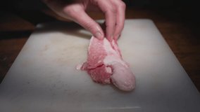 This close up video shows a side view of chef hand slicing fresh pork belly bacon meat with a knife on a cutting board.