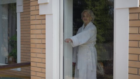 Positive smiling woman opening glass door stretching admiring beauty of sunny day outdoors. Portrait of happy Caucasian mature lady standing on front yard porch in the morning