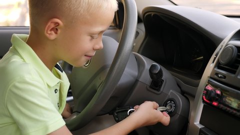 A little cheerful boy sits behind the wheel of a car and turns the ignition key, he pretends to be the driver.
