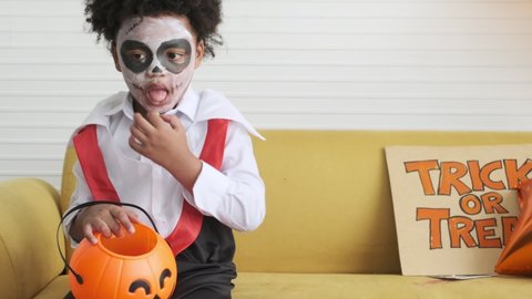 Dark skinned boy in dracula costume eating jelly or candy from pumpkin bucket in halloween party.