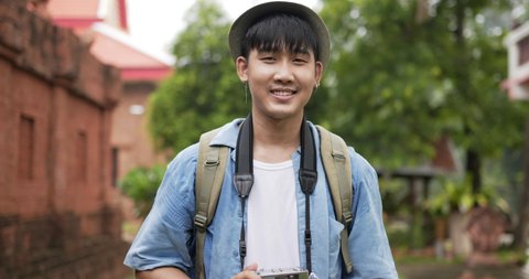 Portrait of Happy asian traveler man with hat taking a photo and looking at camera at ancient temple. Smiling young male walking and holding camera. Holiday, travel and hobby concept.