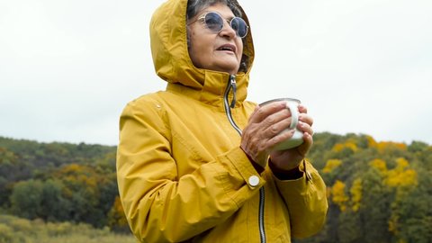 Senior woman rest, relax, outdoor recreation in autumn. Pensioner woman with glasses and yellow raincoat enjoying active leisure and drinking hot beverage from iron mug, outdoors