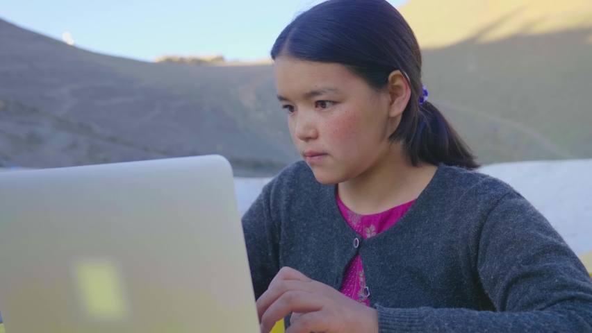 A close shot of a young East Asian focused teenage girl is working on her school project or an assignment wearing a woolen Sweater is typing on a Laptop in a mountainous cold region.  Royalty-Free Stock Footage #1081503296