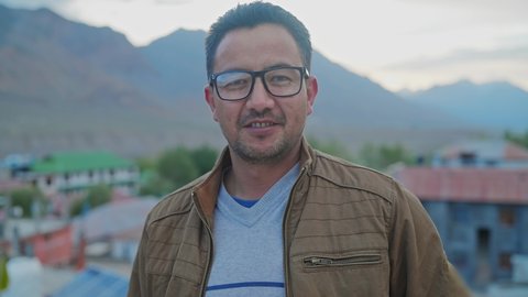 An Asian middle-aged man wearing rimmed Spectacles or eyeglasses and a Leather Jacket standing outdoors in a remote mountainous rural village and looking at the camera with a smile on his face