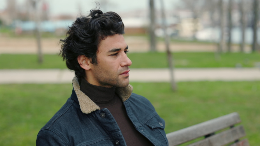 Smiling young guy with dark wavy hair sitting on wooden bench outdoors and looking at camera. Portrait of arabian man sitting alone at city park. Royalty-Free Stock Footage #1081503371