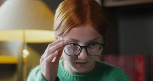 A beautiful red-haired girl in glasses starts a video chat on a laptop, sitting on a sofa in a cozy home interior, close-up. Active facial expressions of a young face