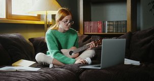 Beautiful red-haired girl wearing glasses dressed in casual clothes learning to play ukulele, while watching video tutorial on laptop sitting on sofa in cozy home interior