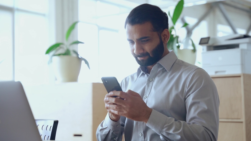 Happy indian ethnic business man entrepreneur investor holding smartphone using mobile app looking at cell phone working with online digital financial data management technology working in office. | Shutterstock HD Video #1081515653