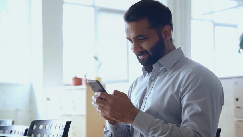 Happy indian ethnic business man entrepreneur investor holding smartphone using mobile app looking at cell phone working with online digital financial data management technology working in office. | Shutterstock HD Video #1081515653