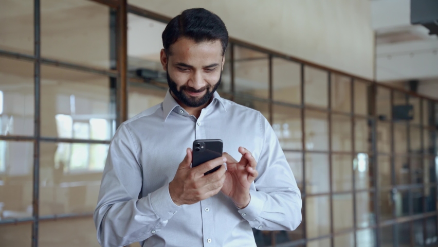 Happy indian ethnic businessman holding smartphone using mobile application program looking at cell phone working checking online digital data market corporate management tech standing in office. Royalty-Free Stock Footage #1081515656