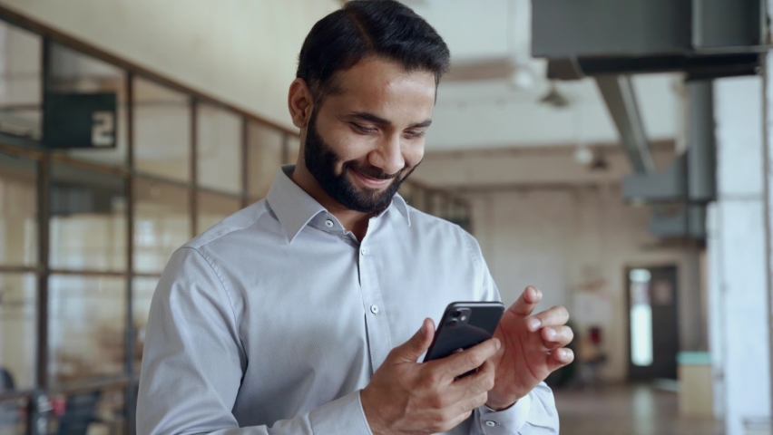 Happy indian ethnic businessman holding smartphone using mobile application program looking at cell phone working checking online digital data market corporate management tech standing in office. | Shutterstock HD Video #1081515656