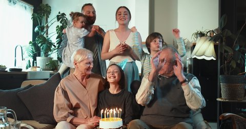 Happy cute teenage girl smiles celebrating birthday blowing on cake under confetti surrounded by cheerful family at home