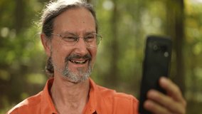 Portrait of ethnic, mature man smiling and laughing while talking to someone on video chat on smartphone with green background outdoors.