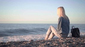 girl is sitting on seashore and dreaming. Romantic walk along beautiful shore. waves . beautiful slender blonde in denim jacket with leather backpack sits on shore and looks at sunset, slow motion