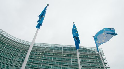 Establishing shot of three waving EU Flags near European Commission Office Building in Brussels, Belgium. Political, Financial or International Business Concept. 4K medium wide orbit shot