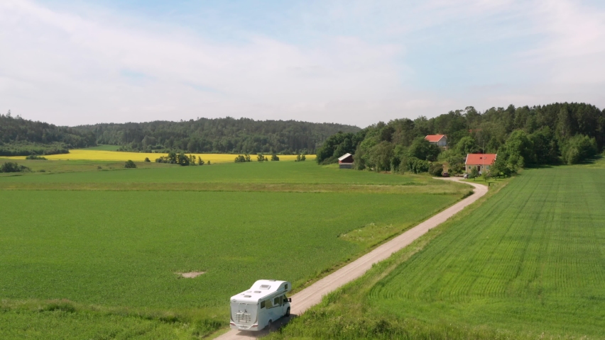 Motorhome caravan driving on gravel country road in Sweden landscape nature. Summer fields and yellow canola rapeseed field and forest. Swedish wooden houses on farmland. Aerial drone shot Royalty-Free Stock Footage #1081524266