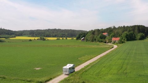 Motorhome caravan driving on gravel country road in Sweden landscape nature. Summer fields and yellow canola rapeseed field and forest. Swedish wooden houses on farmland. Aerial drone shot