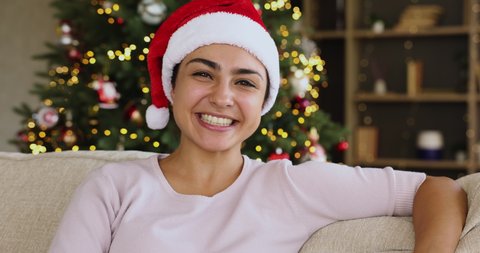 Merry Xmas. Cute laughing Indian woman in Santa hat wave hand to web cam greet friends online. Excited young lady congratulate family with Christmas by videocall speak best wishes near bright fir tree
