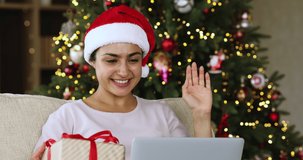 Gifts from Santa. Cheerful young Indian lady in Xmas cap sit on couch chat in video conference app on laptop show gift box to webcam. Excited millennial lady call good friend by pc greet with New Year