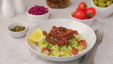 Warm salad with octopus, potatoes, tomatoes, red cabbage, olives, capers and lemon on a white plate. Woman dressing salad with olive oil. Traditional greek dish. Close-up, white background.