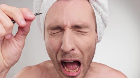 Portrait male looking at mirror, pluck eyebrows and screams in pain. Close up unhappy caucasian man in towel on the head plucking hair between his eyebrows with tweezer. Concept of healthcare