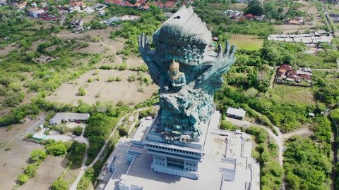 Front rotating aerial view of majestic copper and brass Garuda Wisnu Kencana statue in Bali, Indonesia. Large blue green statue rising above the residential homes